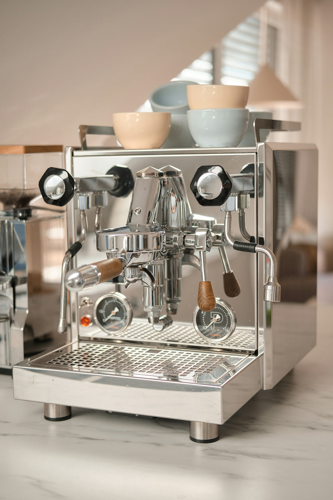 A chrome espresso machine with wooden handles and ceramic cups on a marble counter